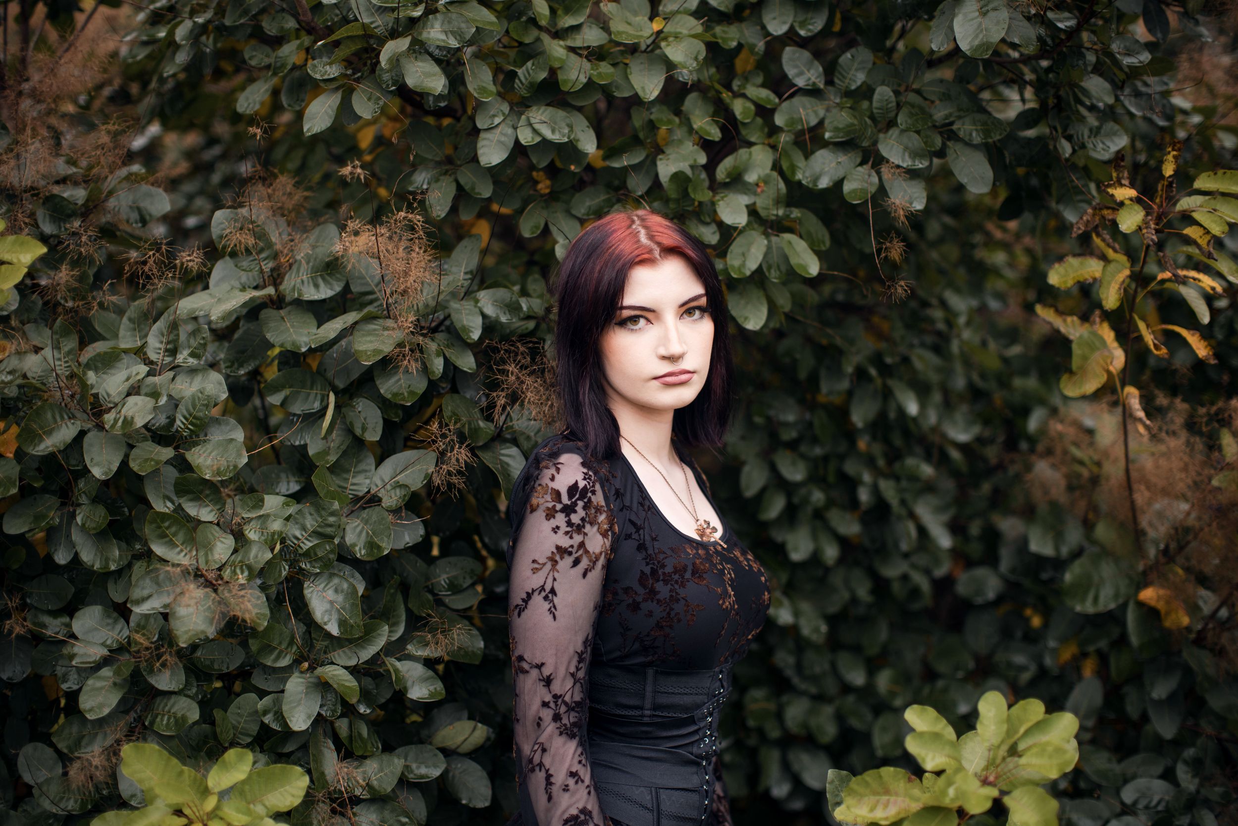 Young woman with red hair in black lace top poses against lush green ivy wall outdoors.