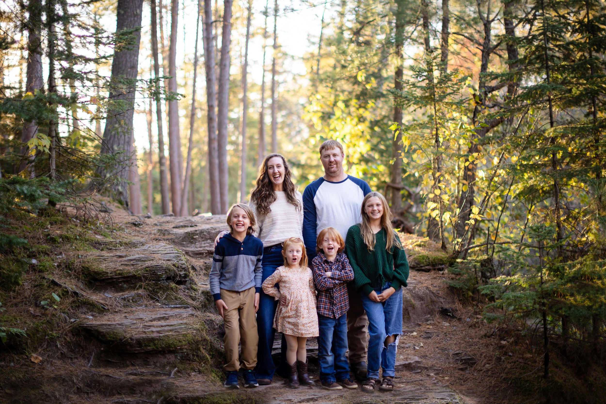 Family of five posing on a forest trail surrounded by tall trees and autumn foliage in a professional outdoor portrait.