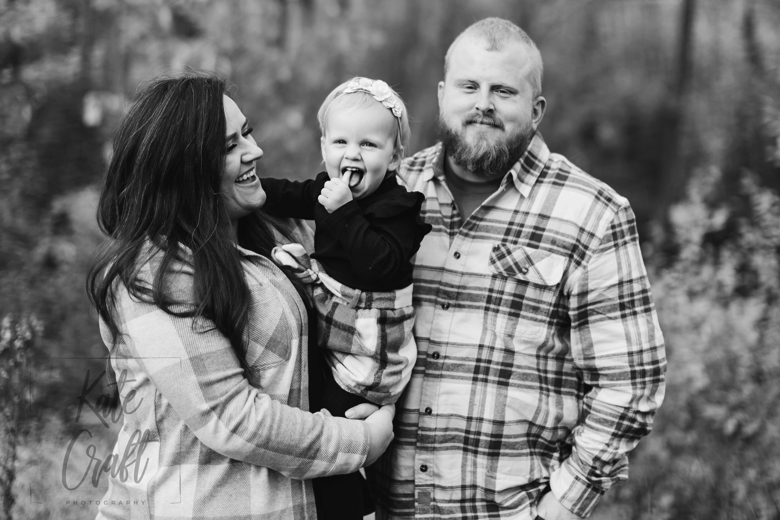Black and white family portrait of smiling parents holding their toddler outdoors among trees.