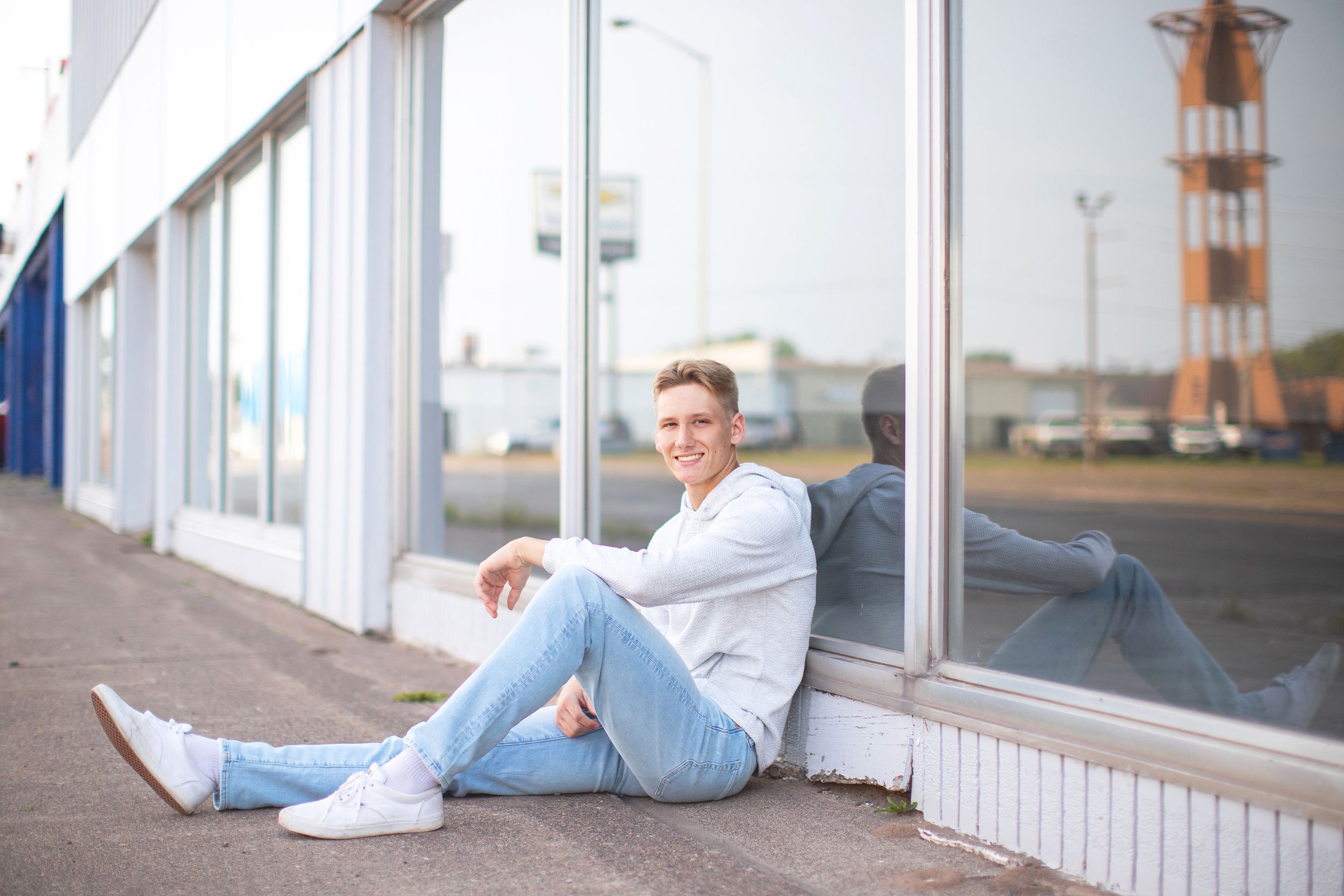 Young man in white sweater and jeans sitting against a storefront window, smiling outdoors on a sunny day.