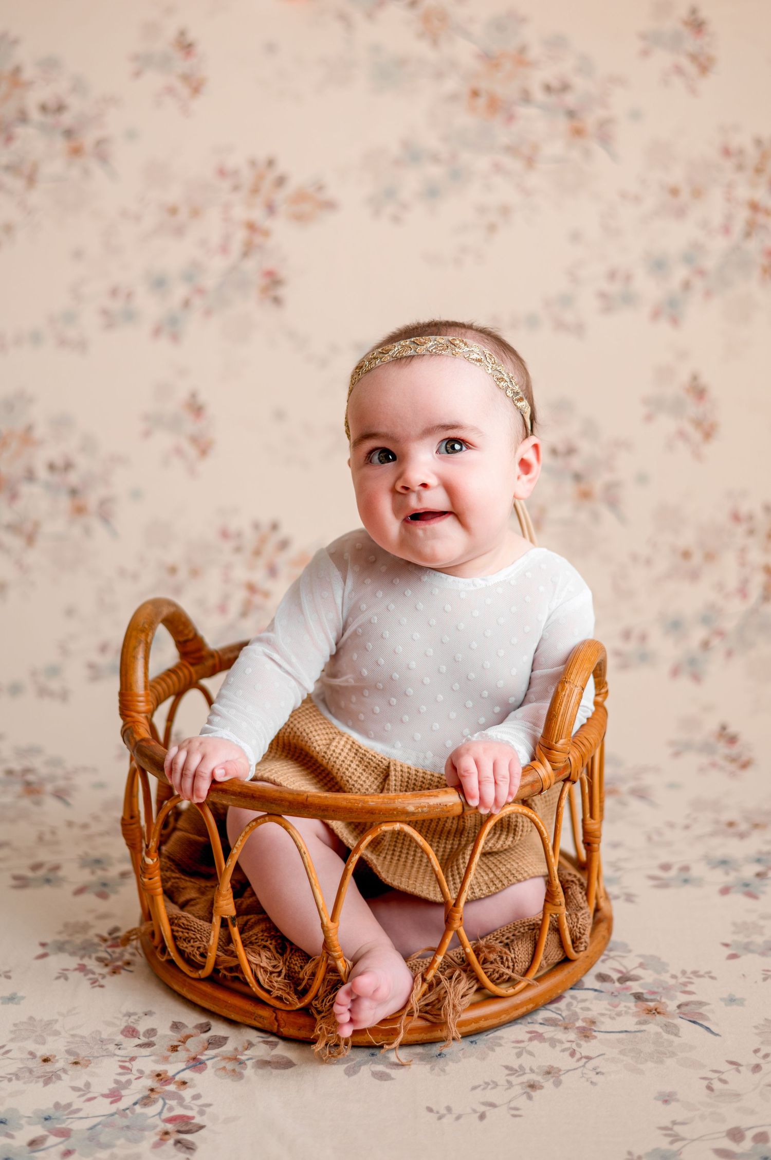 Red-haired baby in beige knit romper sitting on small wooden chair against green background.