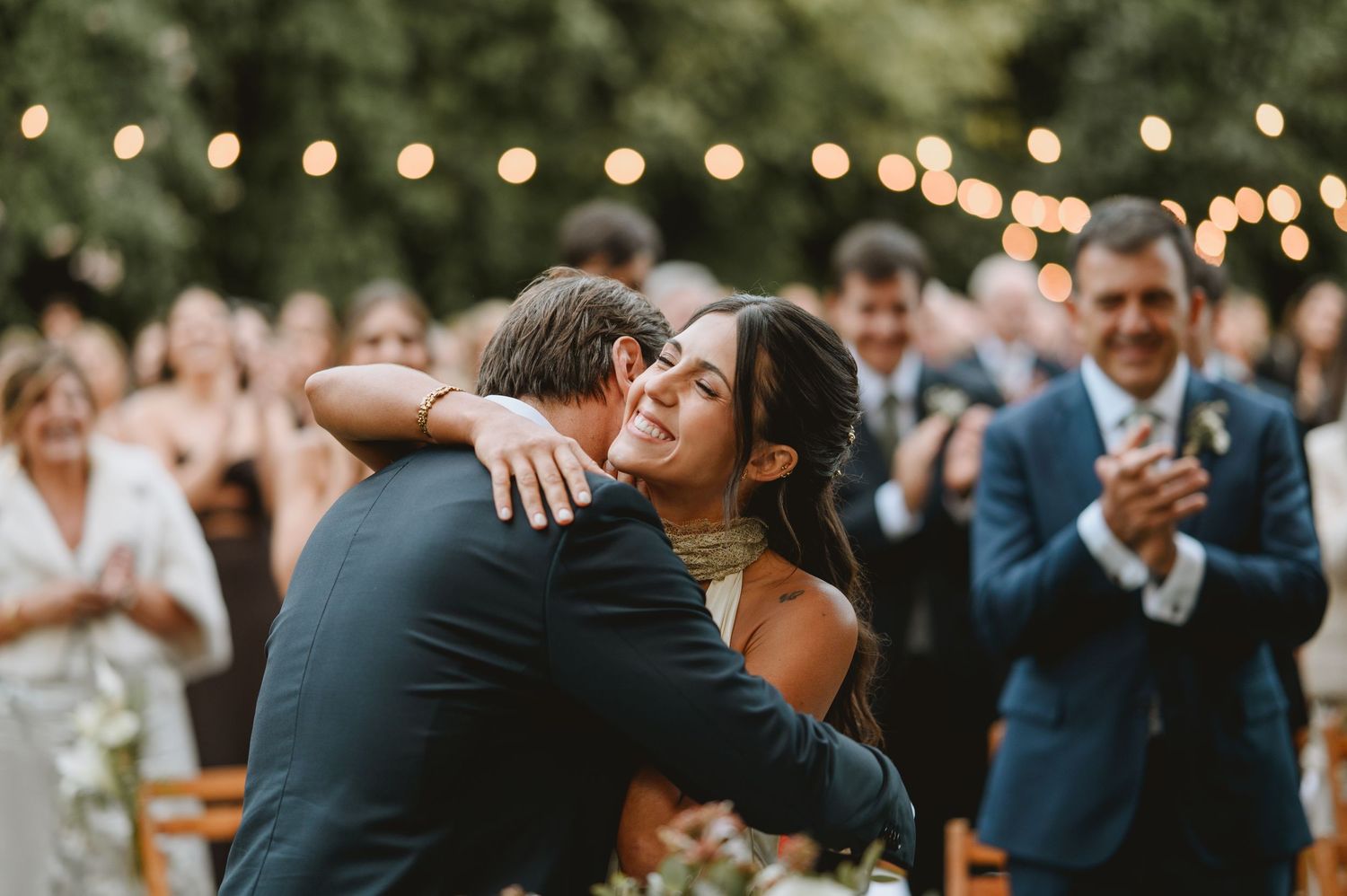 Bride in white halter gown sharing emotional hug with groom at outdoor wedding with string lights above.
