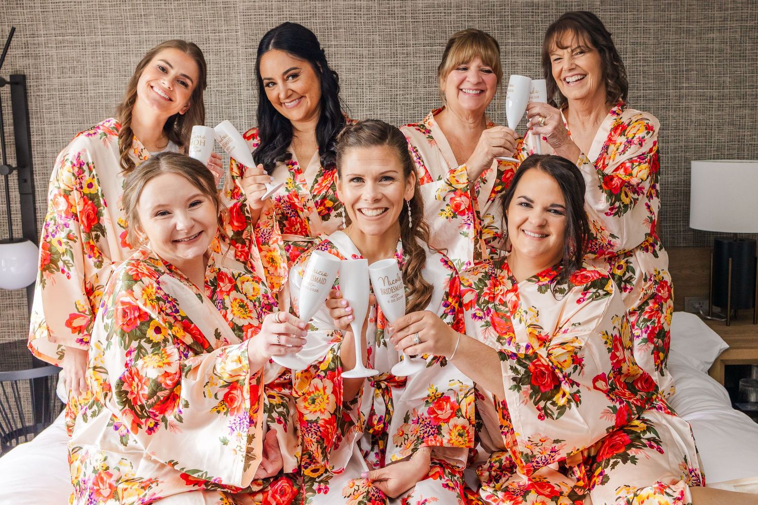 Seven bridesmaids in matching floral robes smile and toast with champagne flutes during a bridal party getting-ready celebration.