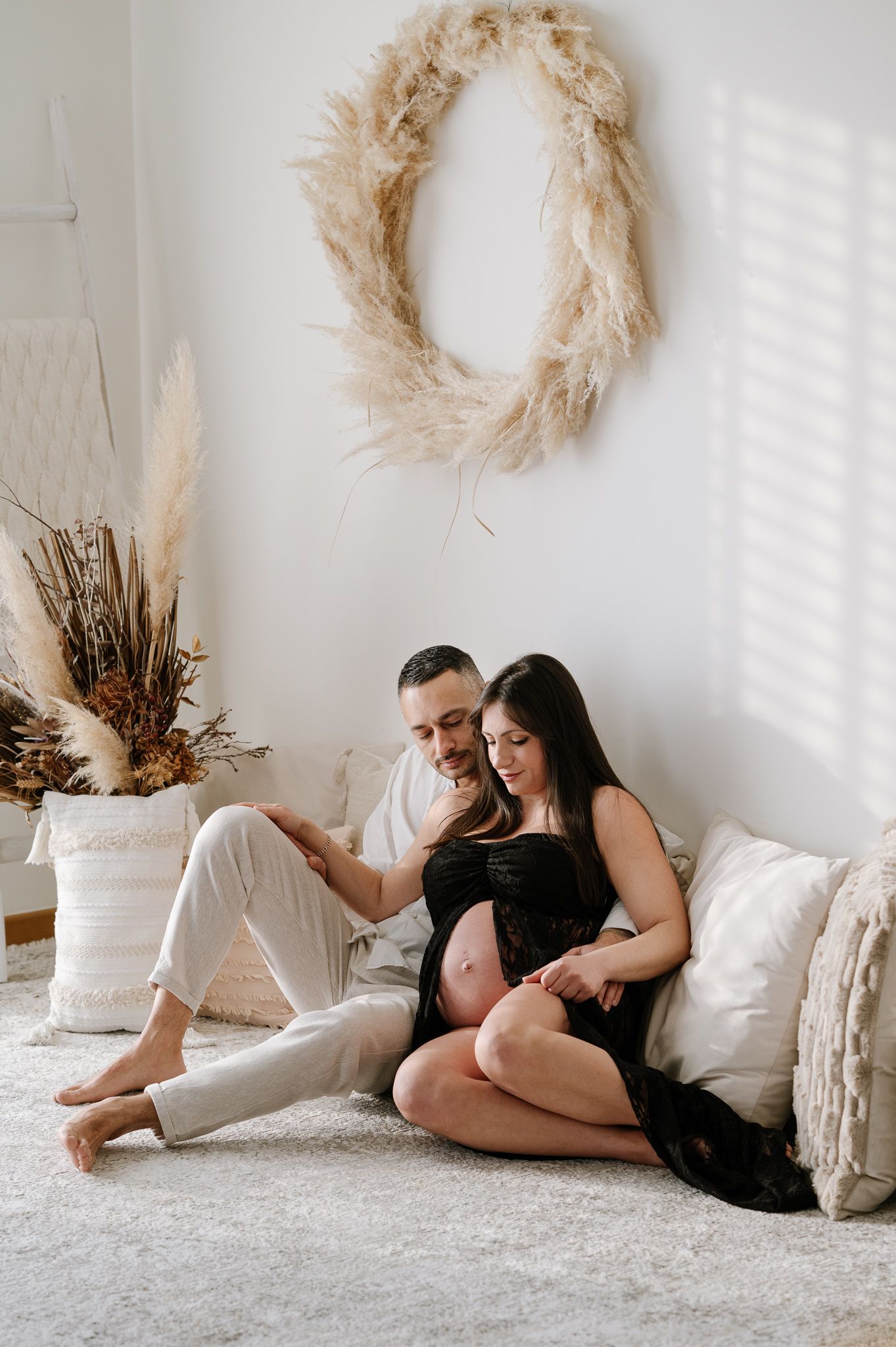 Couple sitting on floor during maternity photoshoot, woman in black dress showing baby bump, boho decor with pampas grass wreath.