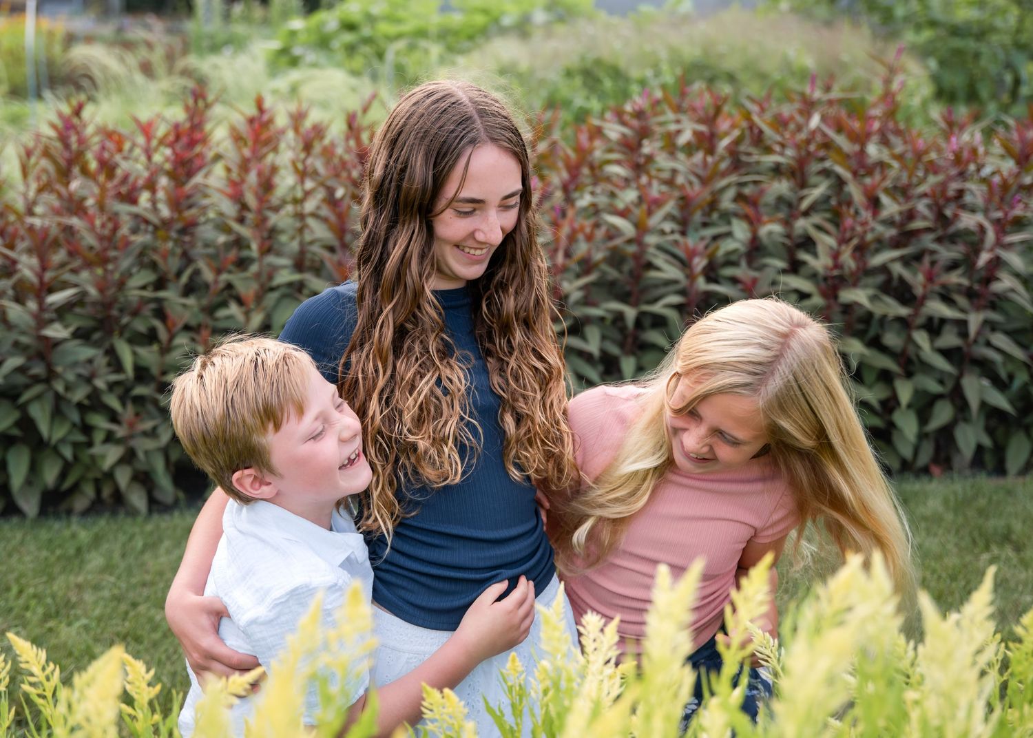 Two girls and a young boy laughing together in a lush green garden with colorful plants.