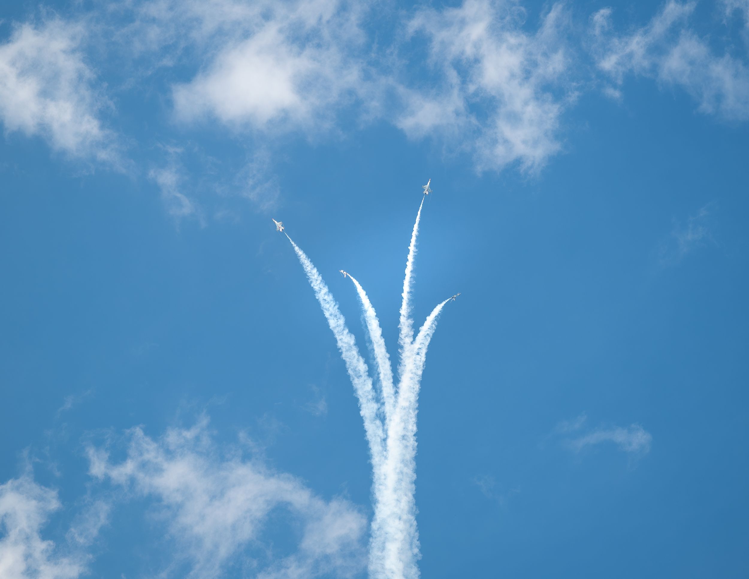 Four jets perform a dramatic aerial maneuver, leaving white vapor trails against a bright blue sky with scattered clouds.
