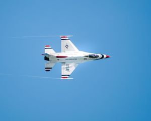 USAF Thunderbirds F-16 jet performing aerial maneuver against clear blue sky, viewed from below showing red, white, and blue markings.