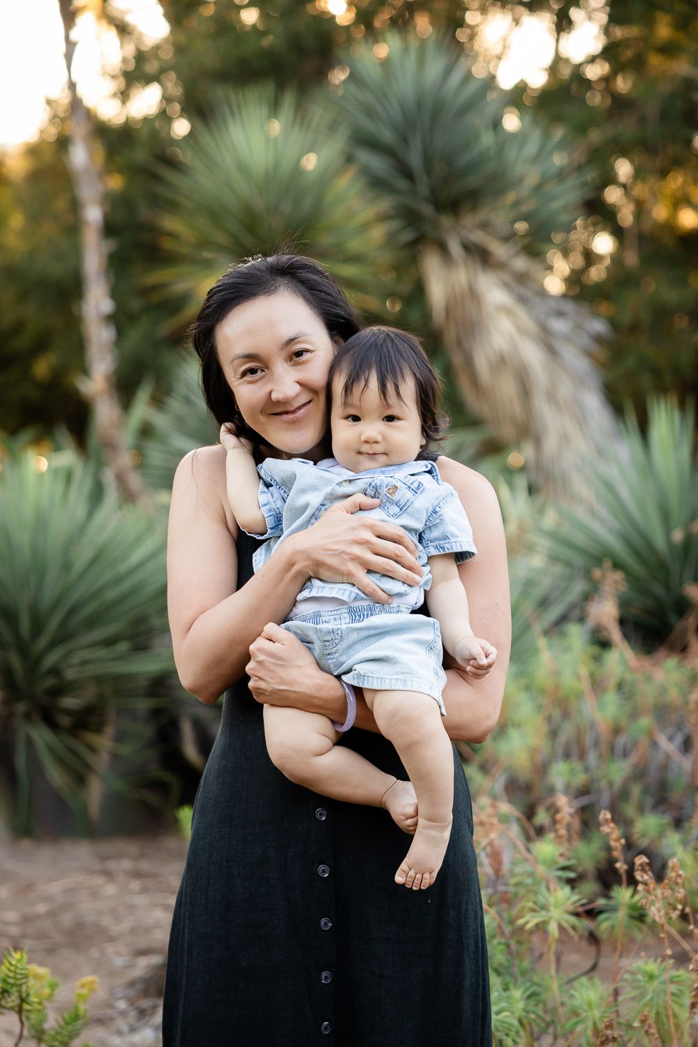 A smiling mother in a black dress holds her baby boy outdoors among lush green plants in warm sunlight in the Bay Area