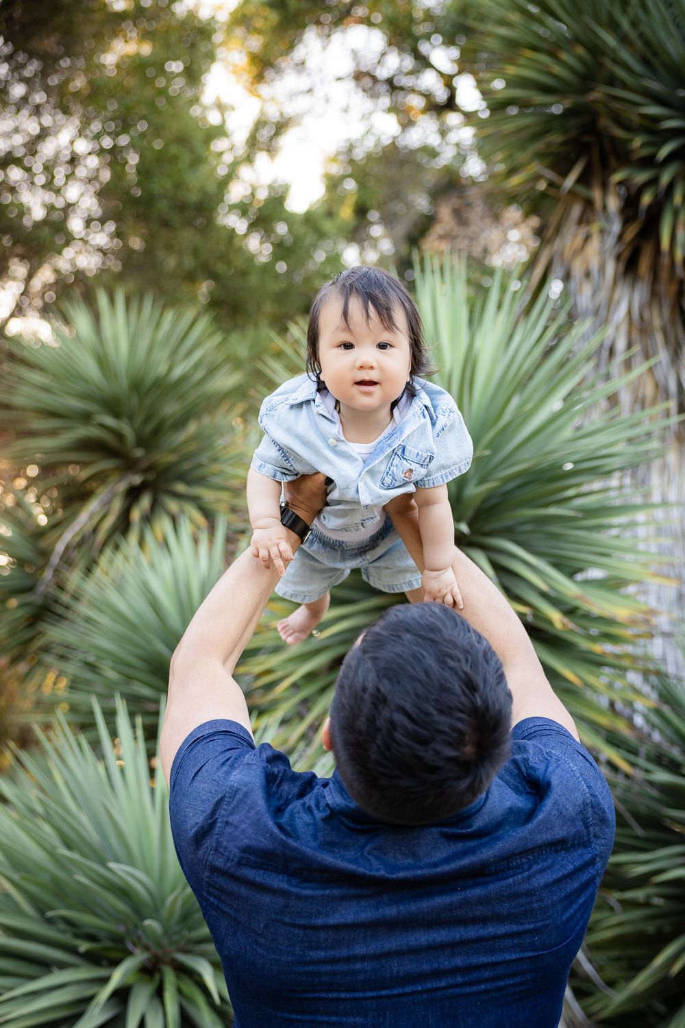 Father lifting happy baby in the air outdoors, surrounded by lush green tropical plants during their outdoor family photoshoot in Palo Alto