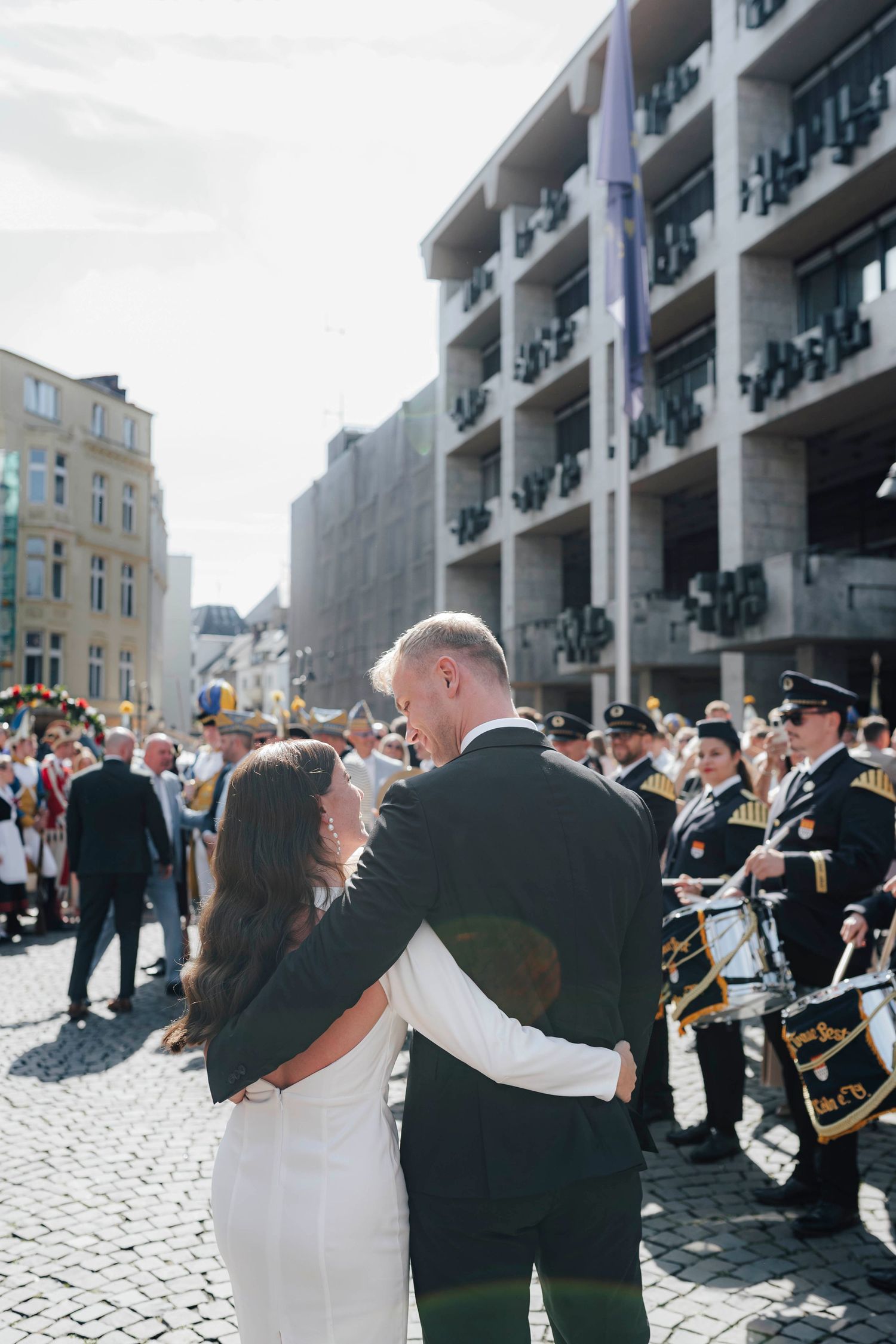 A bride and groom embrace on a cobblestone street with a marching band and crowd celebrating their wedding outdoors.