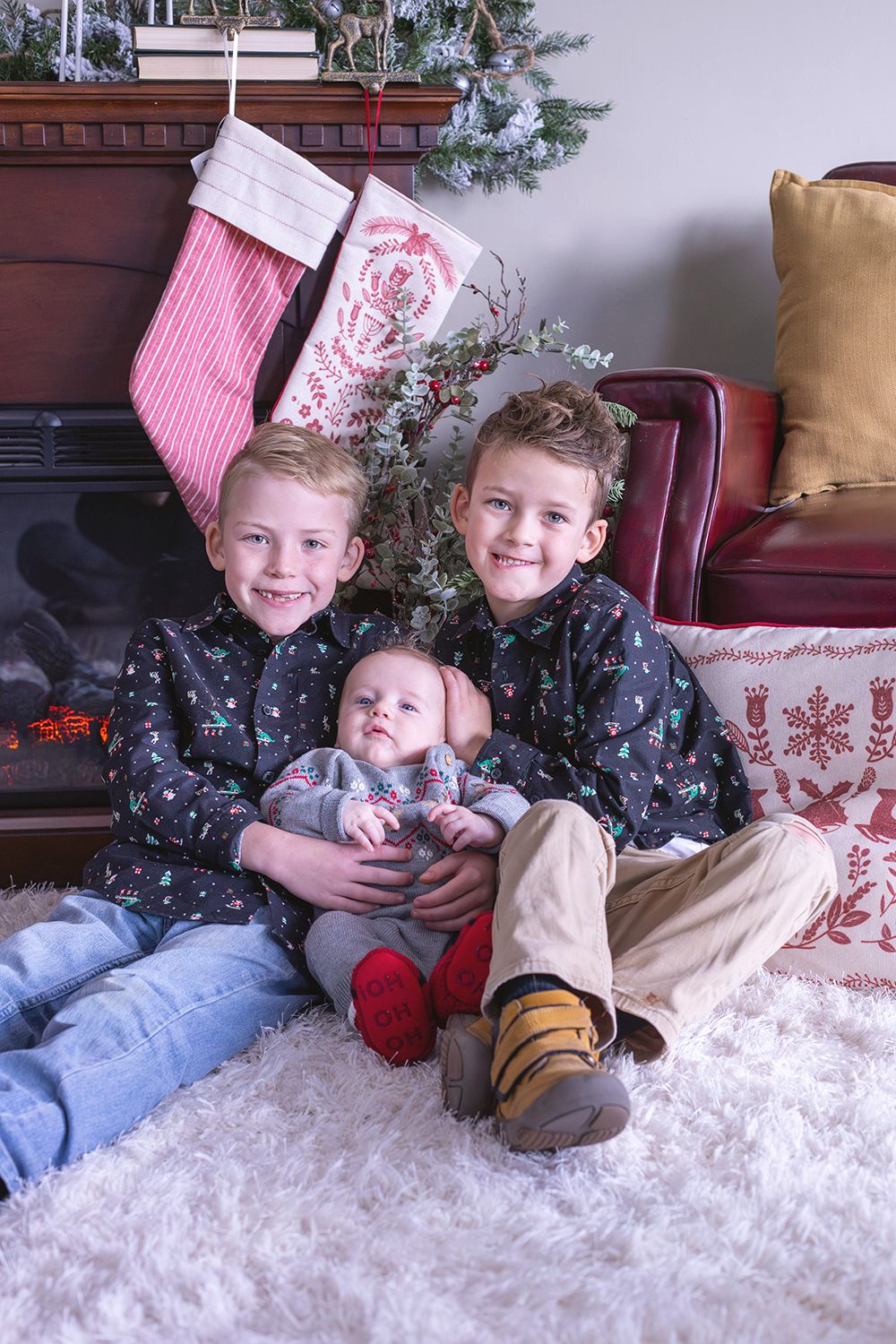 Two boys in matching Christmas shirts hold a baby sibling on a white rug by a festive fireplace with stockings.