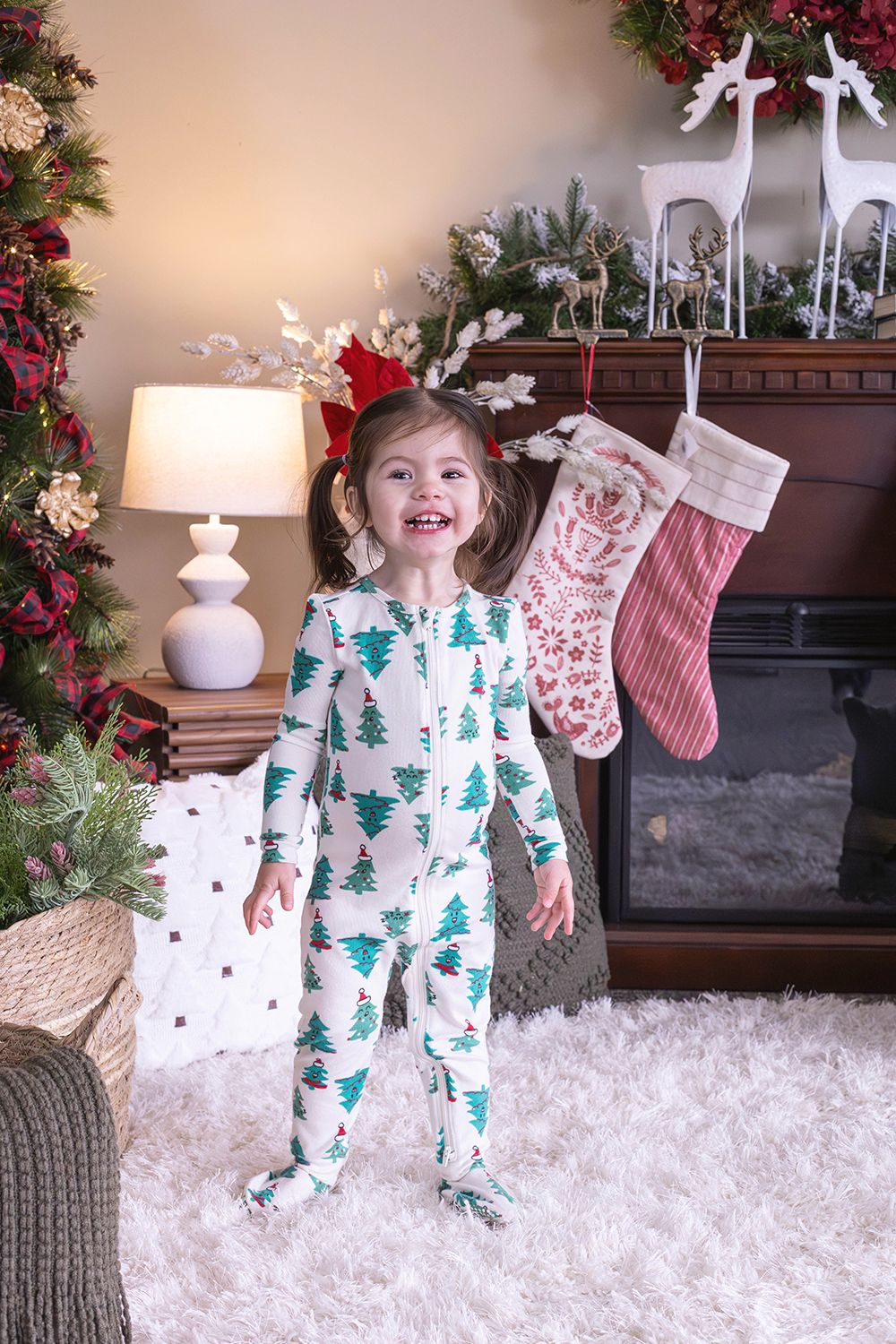 Smiling toddler girl in Christmas tree pajamas stands by decorated fireplace with stockings and holiday decor.
