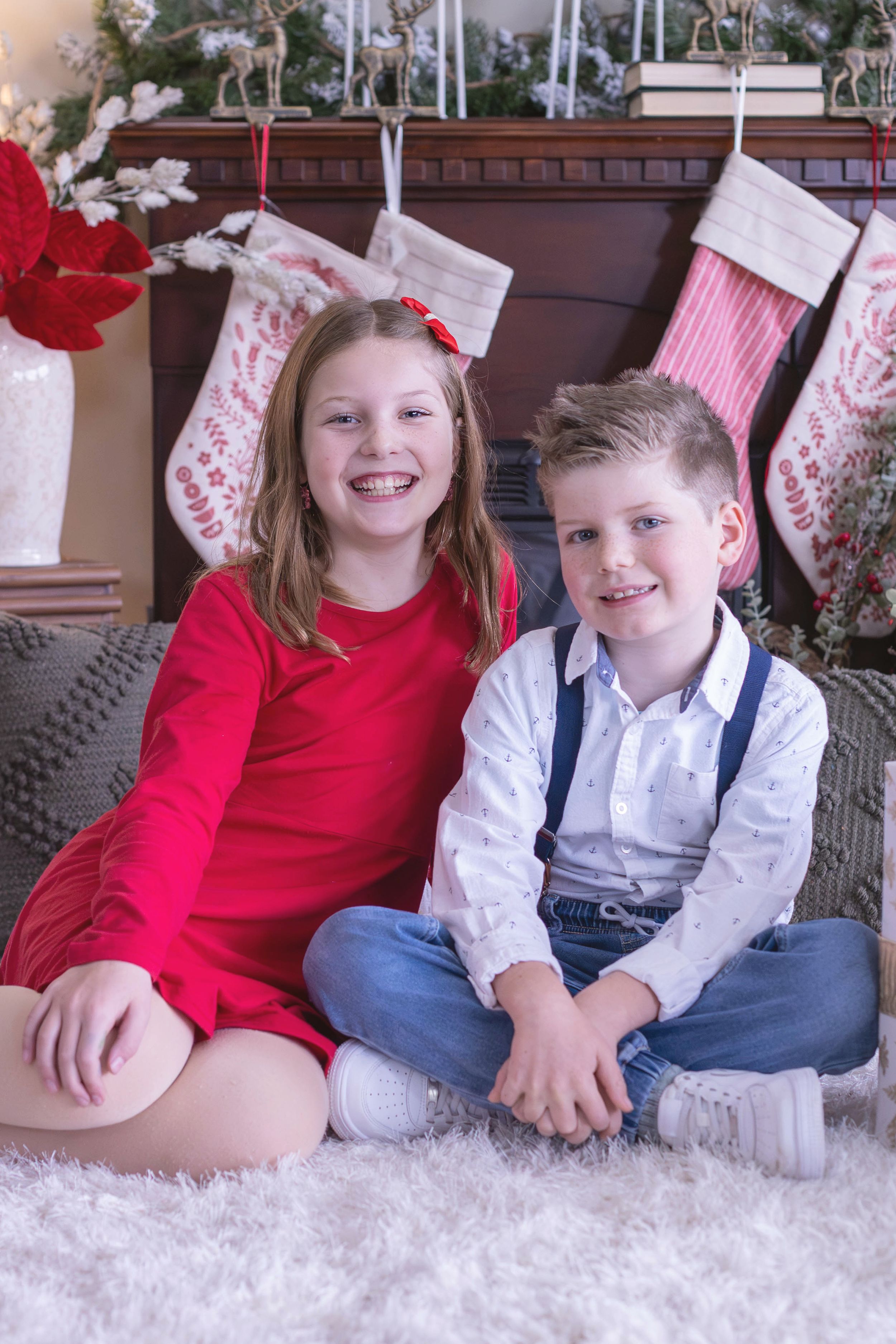 Two smiling children in holiday outfits sit on a fluffy rug in front of a Christmas fireplace with stockings.