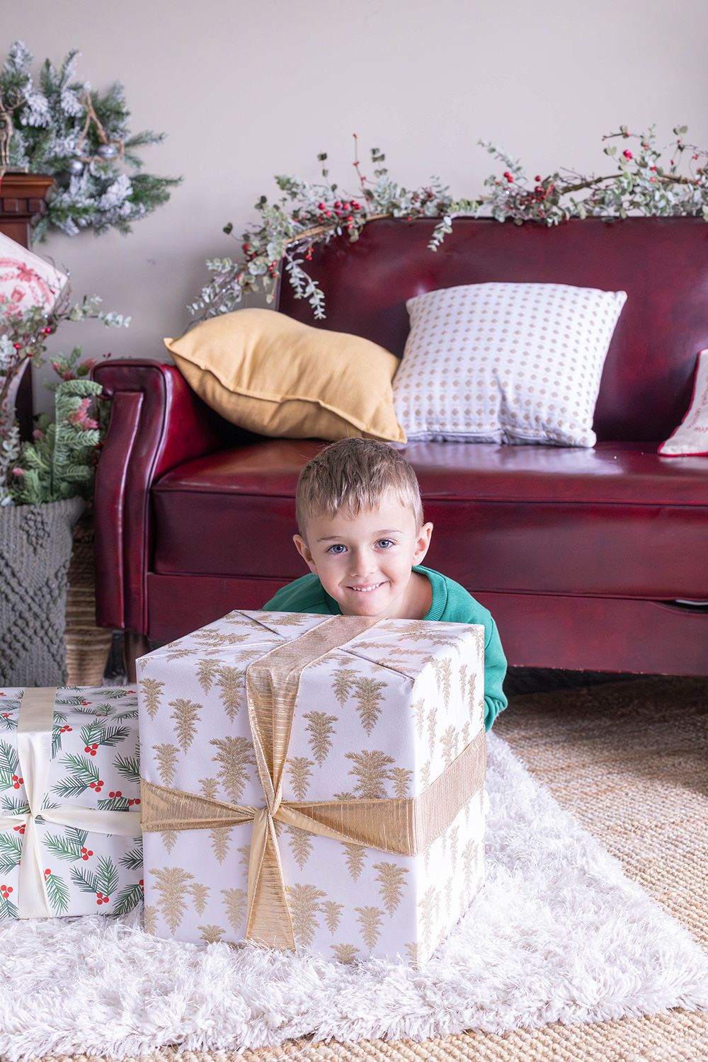 Smiling boy lying on white rug with large Christmas gift, red leather sofa with pillows and holiday decorations behind him.