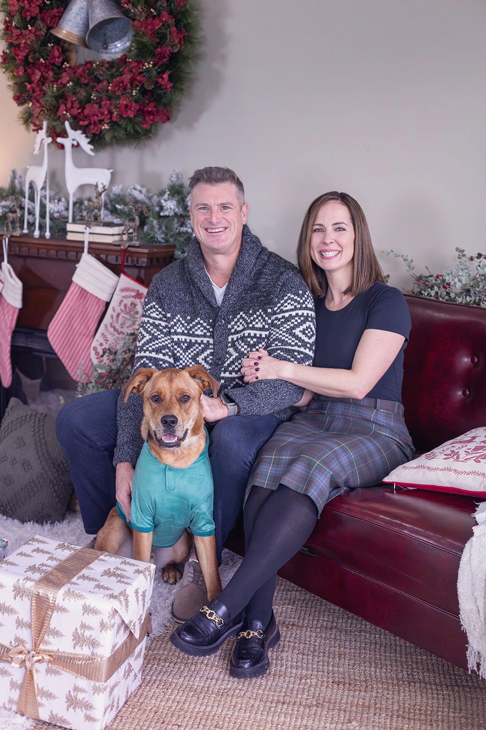 Couple smiling on red sofa with dog in teal shirt, Christmas wreath, stockings, and gifts in festive holiday setting.