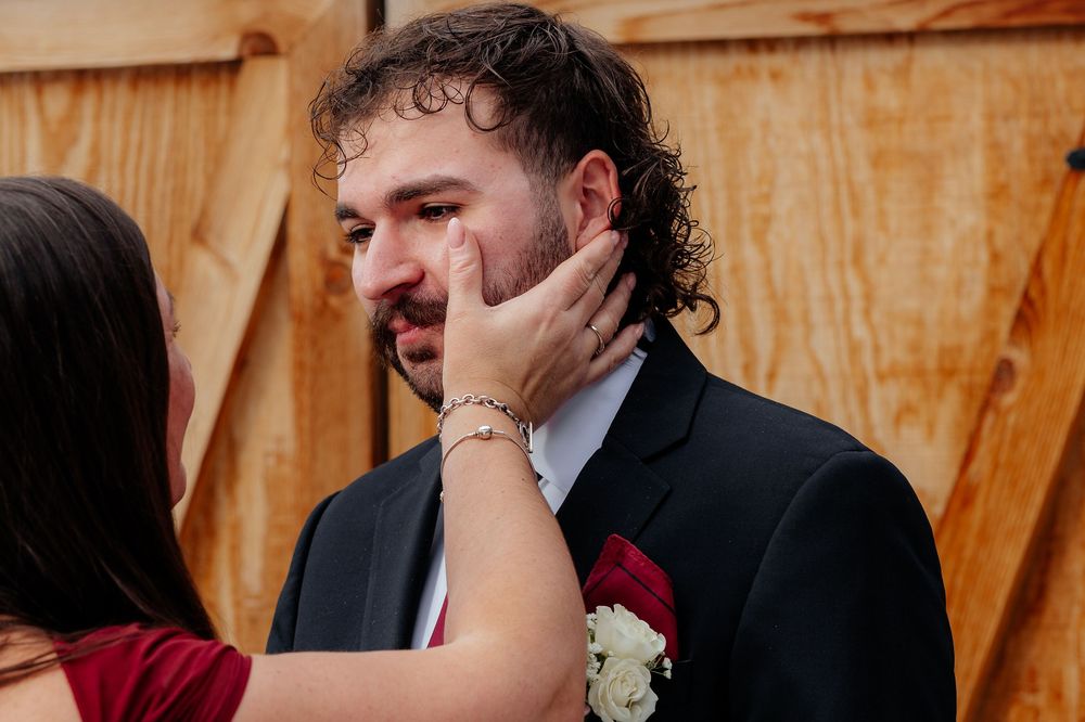 A groom in a dark suit with a red boutonniere has his face tenderly cupped by a woman's hands at a wedding.