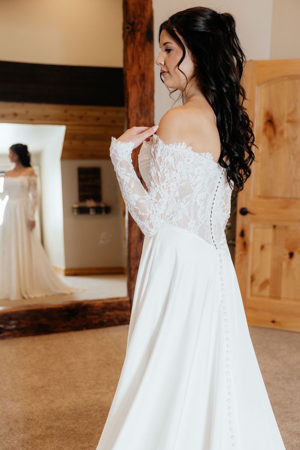 Bride in off-shoulder lace wedding gown with long sleeves, viewed from behind near mirror.