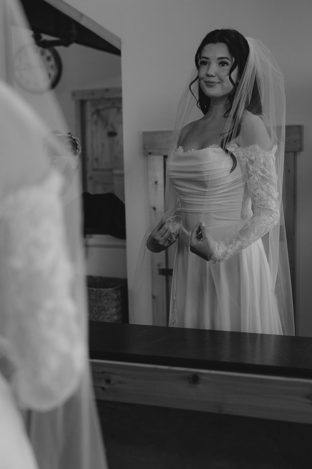 Black and white photo of a bride in a lace wedding dress and veil admiring her reflection in a mirror.
