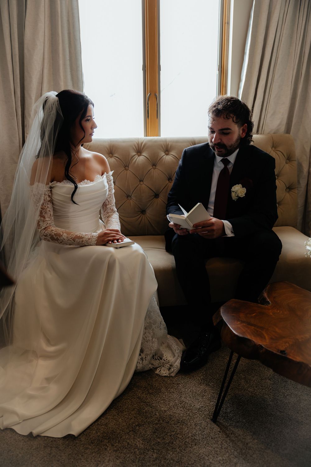 Bride and groom sitting on a tufted sofa exchanging private vows, with the groom reading from a small notebook.
