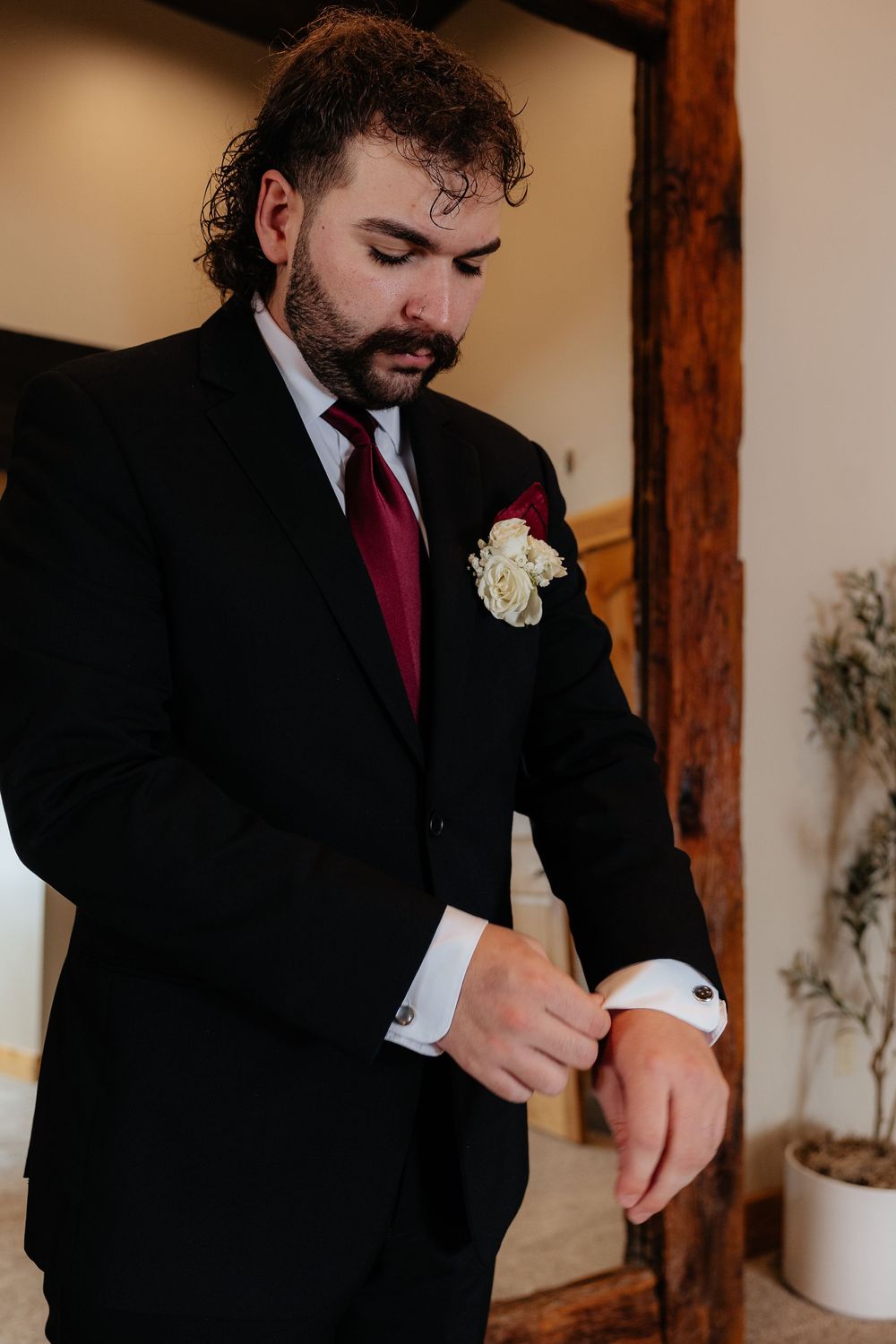 Groom in black suit with burgundy tie and white boutonniere adjusting cufflinks before wedding ceremony.