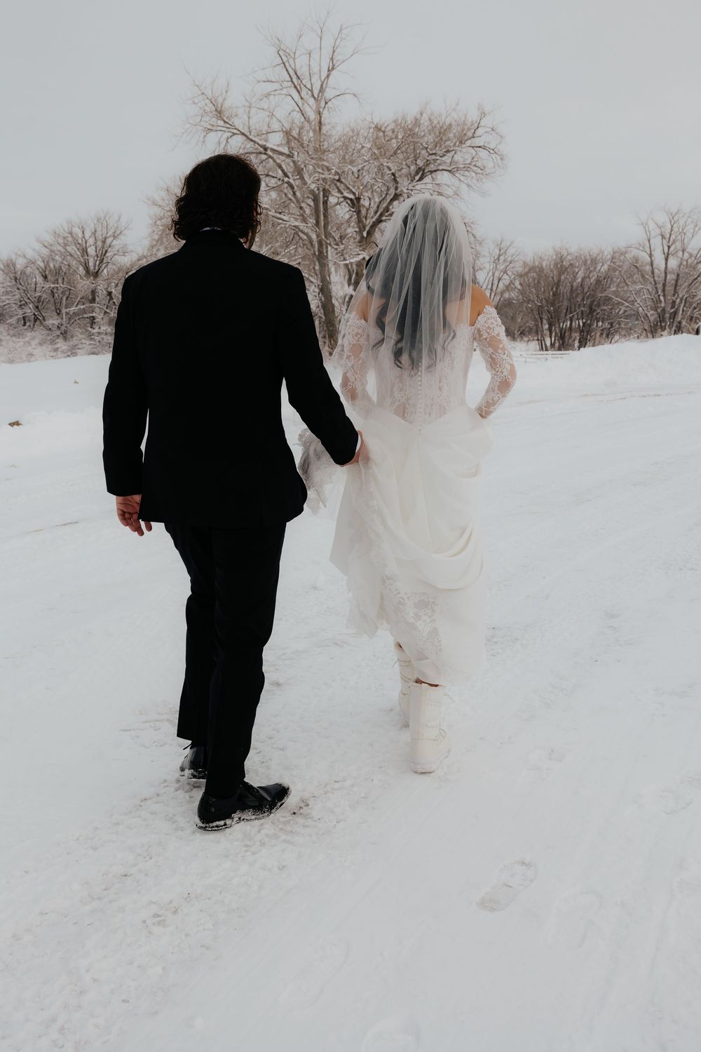 Bride and groom walking hand in hand through snowy winter landscape, bride wearing white gown with veil and groom in black suit.