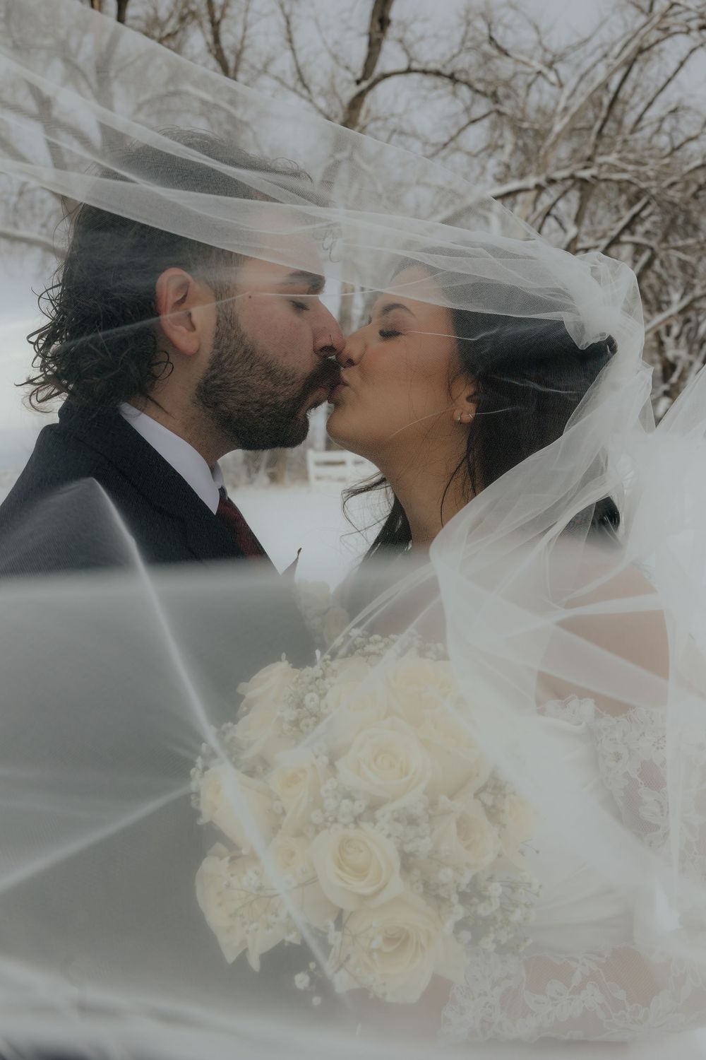 Newlyweds kissing under flowing veil in snowy winter setting, bride holding white rose bouquet.
