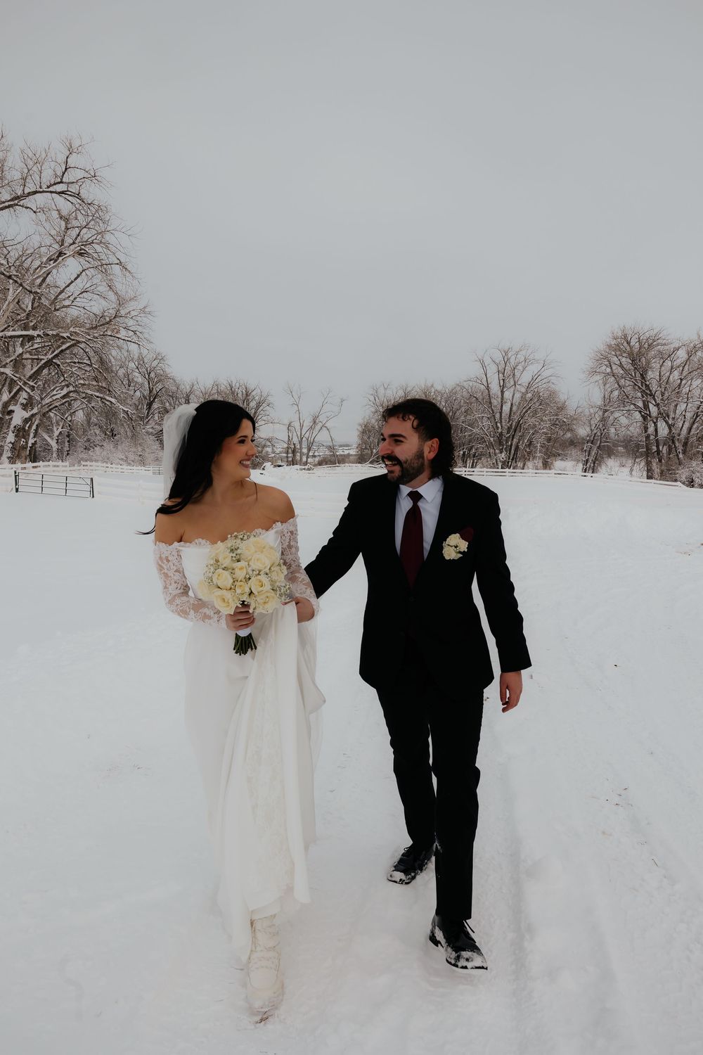 Bride and groom walk hand-in-hand through snowy winter landscape, bride holding white bouquet in lace off-shoulder gown.