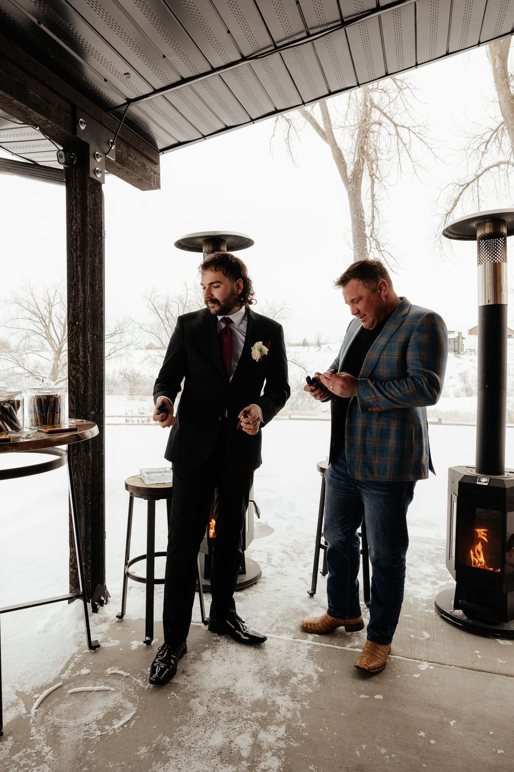 Groom in black suit with boutonniere stands beside man in plaid blazer near patio heater at winter wedding venue.