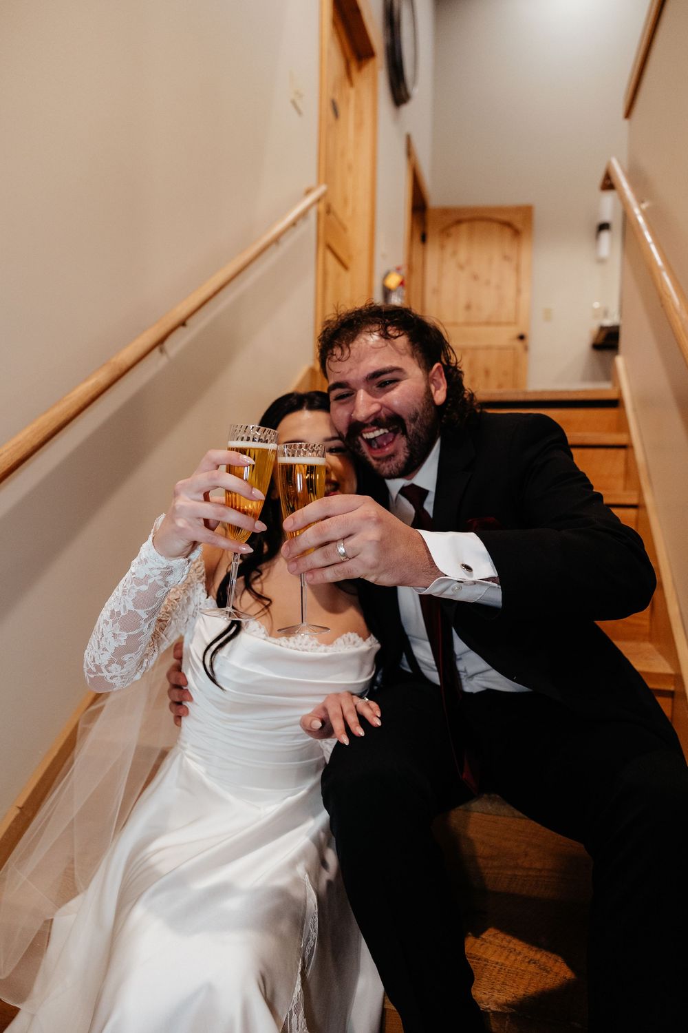 Bride and groom laughing and toasting with beer glasses while sitting on wooden stairs at their wedding reception.