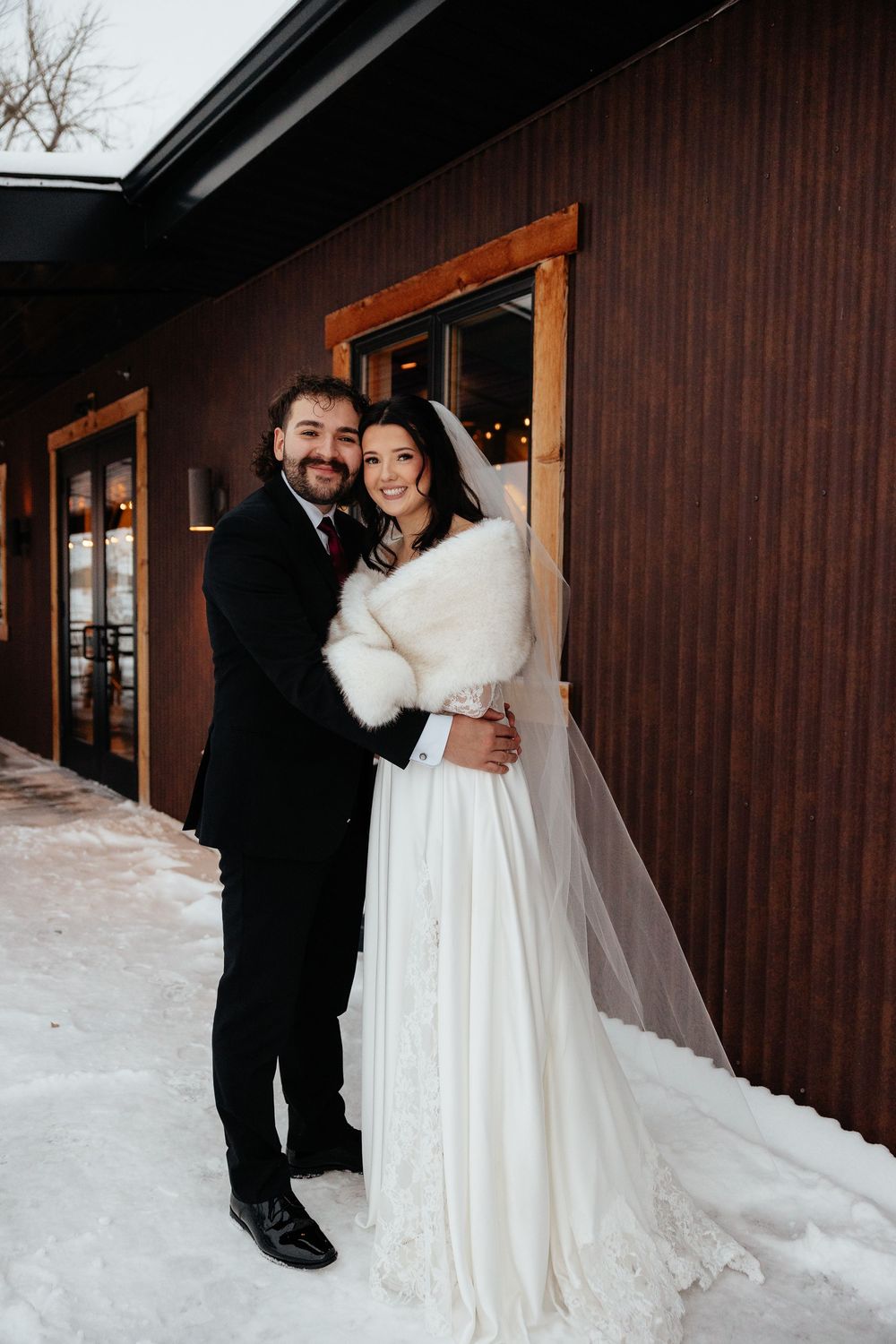 Winter wedding couple embracing outside snowy venue, bride in white gown with fur wrap and veil, groom in black suit.