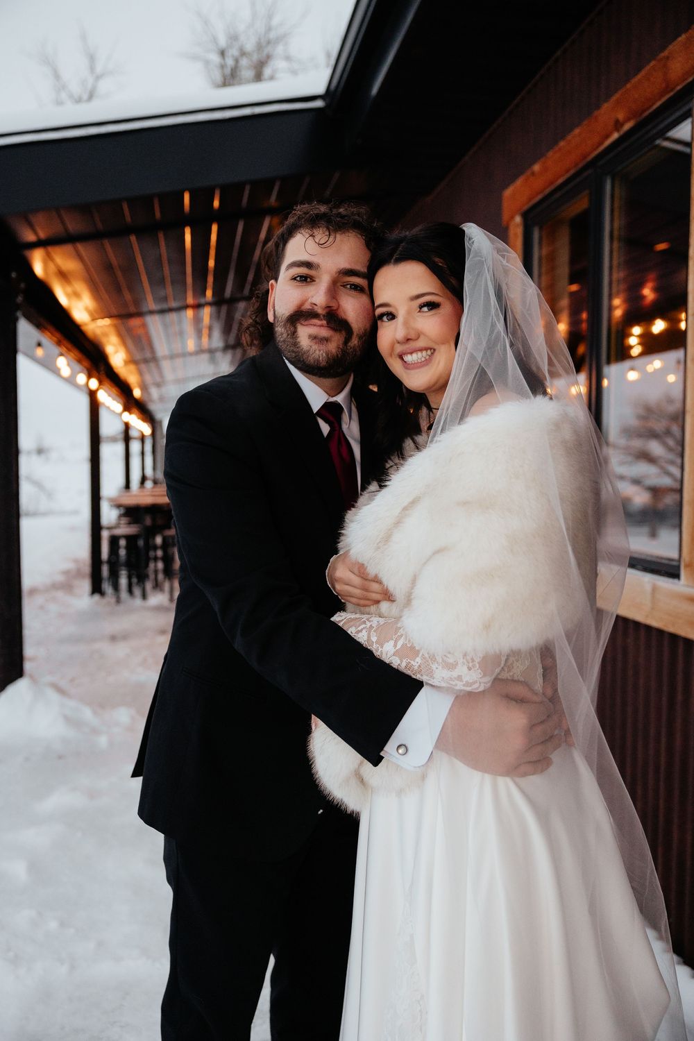 Smiling newlyweds posing outdoors in winter, bride wearing fur stole and veil, groom in dark suit with burgundy tie.