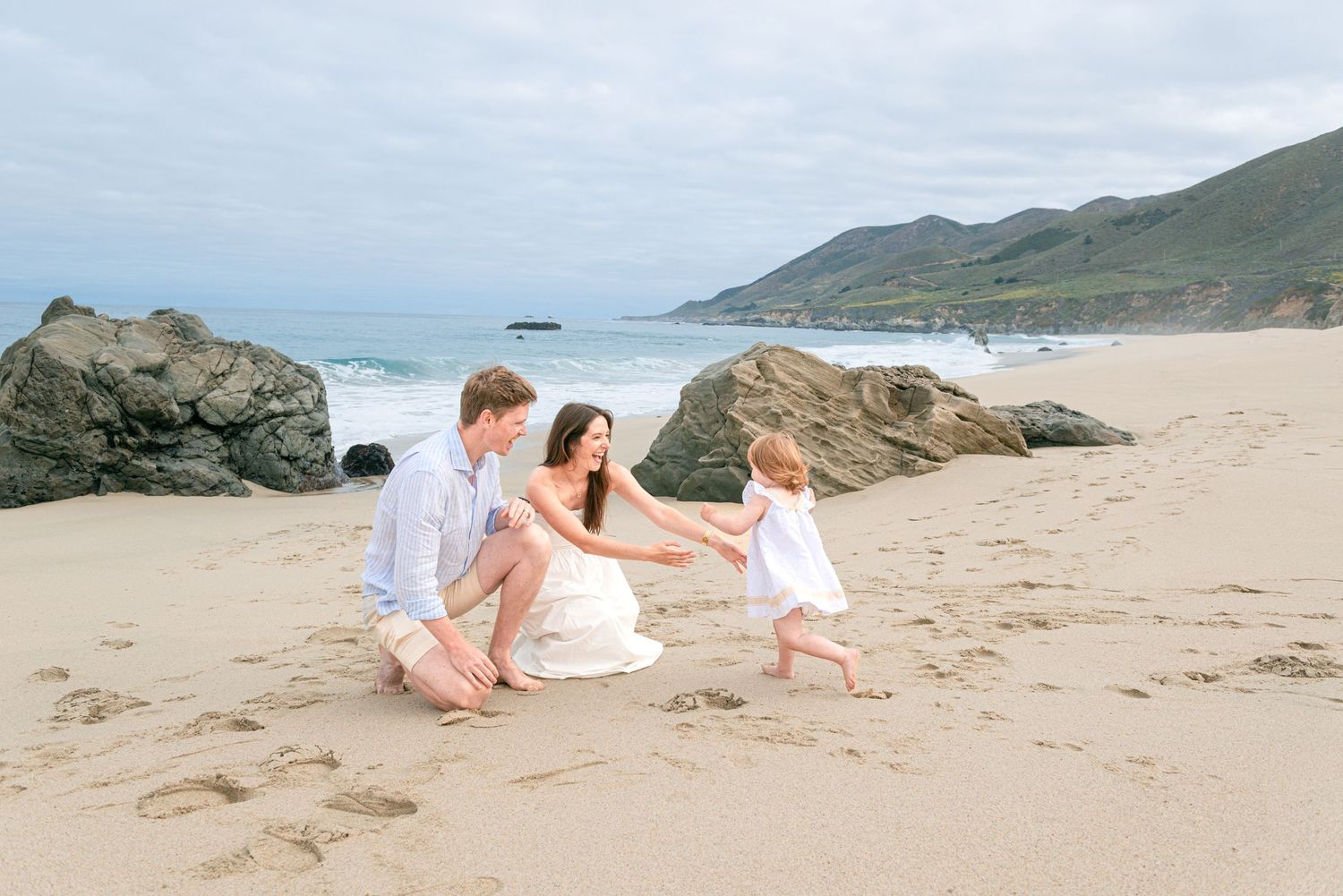 Family of three enjoying a sunny beach day, parents crouching as toddler walks toward them on sandy shore.