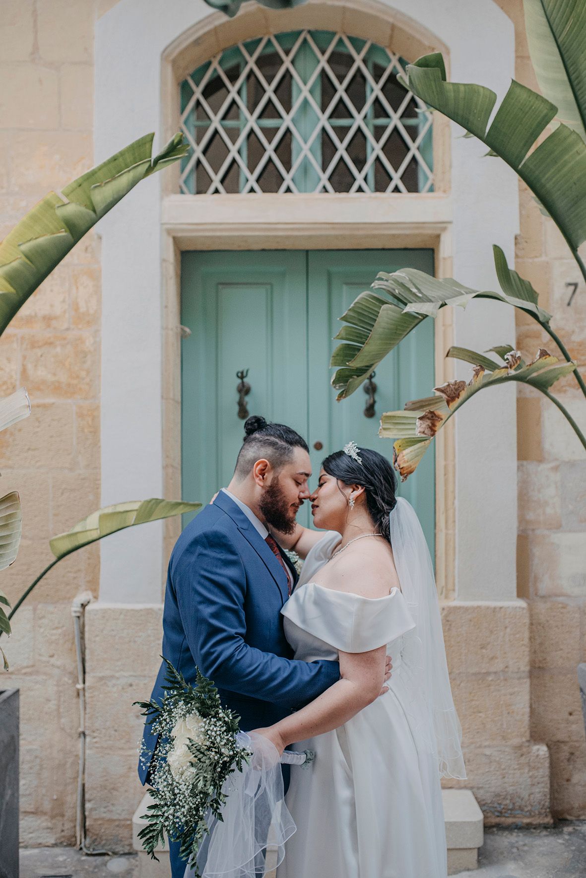 Bride and groom share a romantic moment under a tropical arch before a teal door on their wedding day.