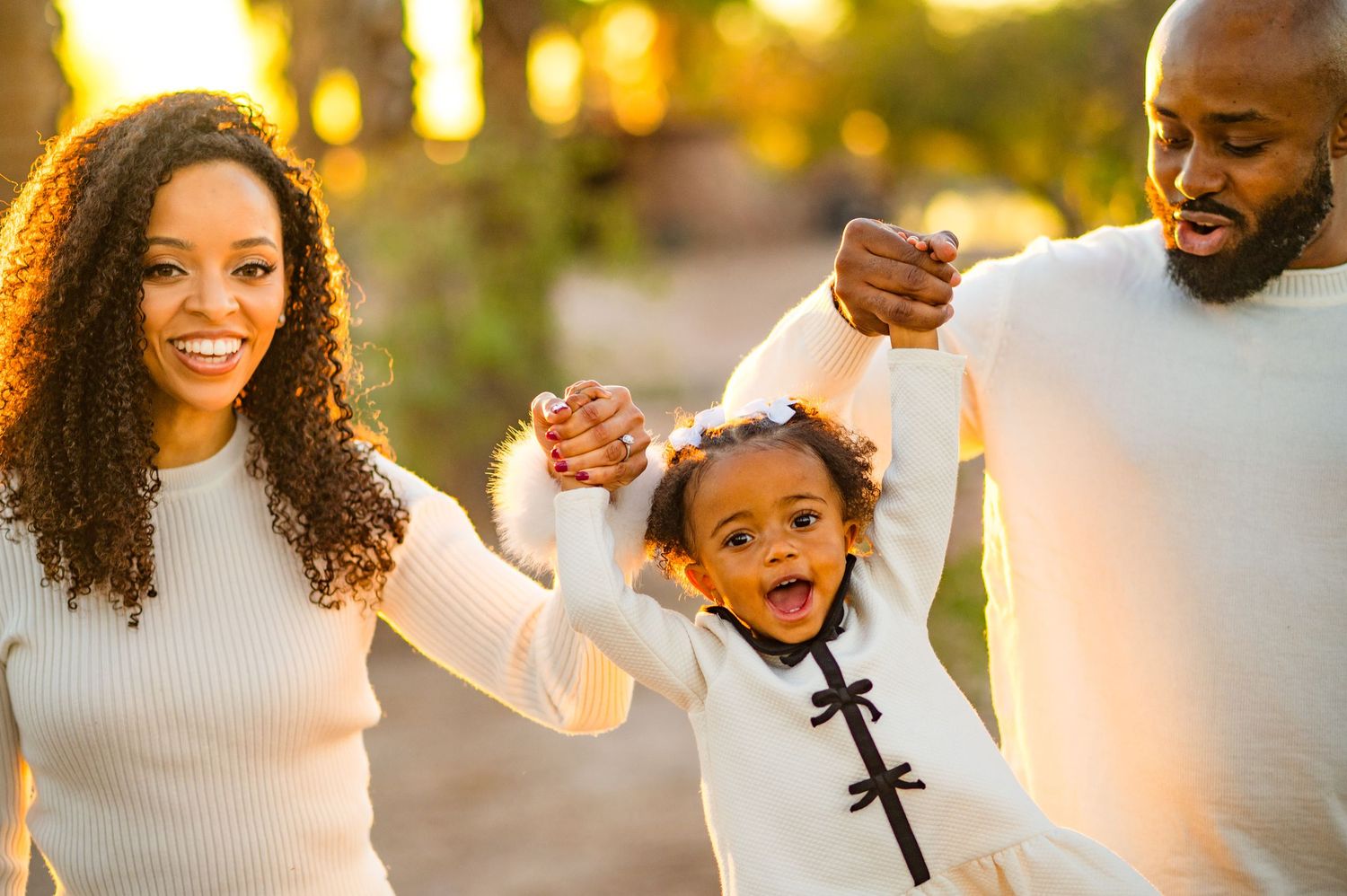 Happy Black family with parents swinging young daughter outdoors in warm golden sunlight, all wearing white outfits.