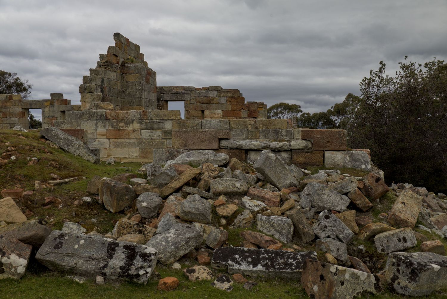 Ruins of an ancient stone building with scattered rocks and rubble under a cloudy sky in a rural landscape.