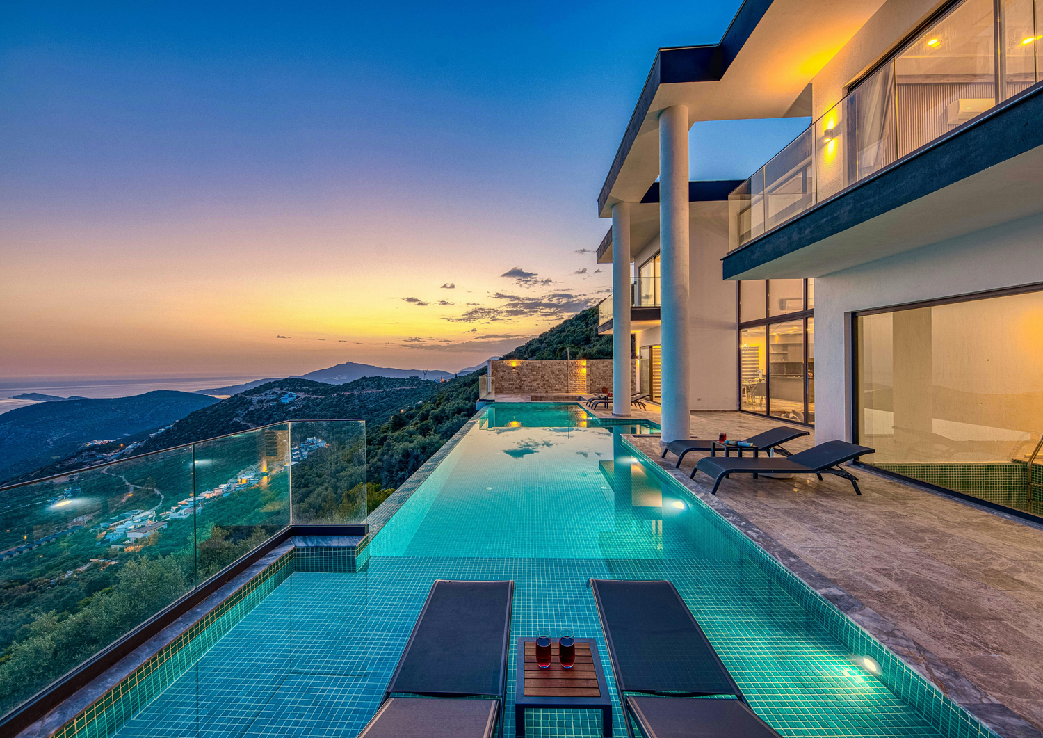 Stunning infinity pool villa at sunset with panoramic ocean views, sun loungers, and illuminated modern architecture.