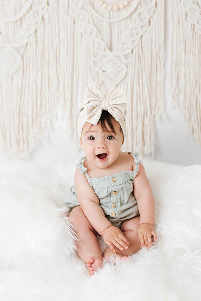 Smiling baby girl in sage romper and cream bow headband sitting on fluffy white rug with macrame backdrop.