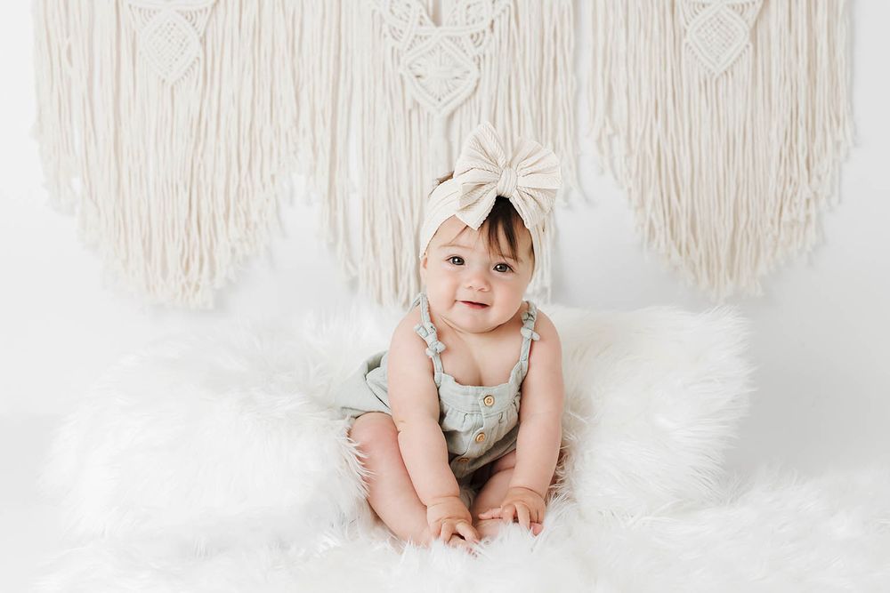 Adorable baby girl in floral romper and cream bow headband sitting on fluffy white rug with macrame backdrop.