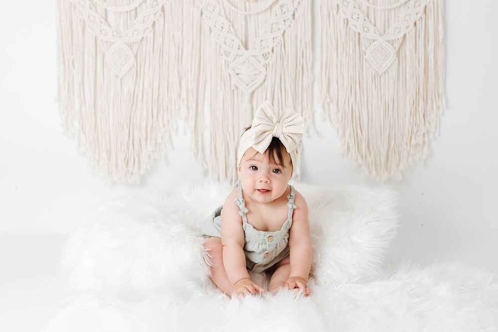 Adorable baby girl in floral romper and bow headband sits on white fur rug before macrame wall hanging.