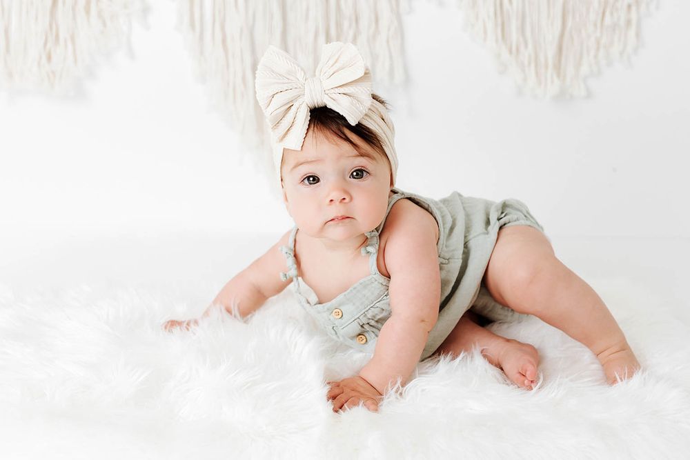 Adorable baby girl in sage green romper and cream bow headband lying on white fur rug.