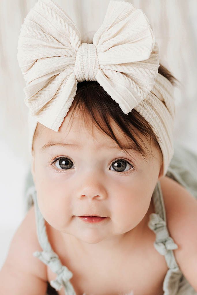 Close-up portrait of a cute baby girl wearing a large cream ribbed bow headband with dark expressive eyes.