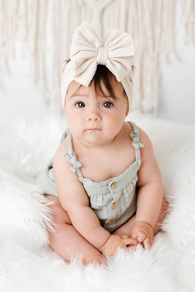Adorable baby girl in sage green romper and cream bow headband sitting on fluffy white rug.