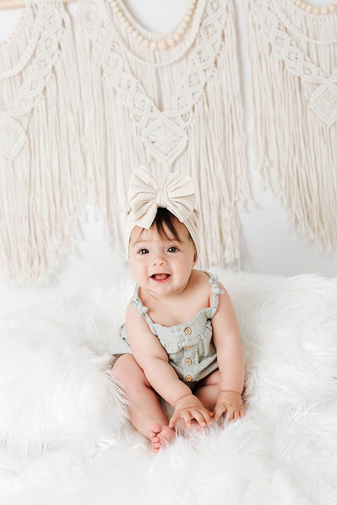 Smiling baby girl in sage romper and bow headband sitting on fluffy white rug with macrame wall hanging.