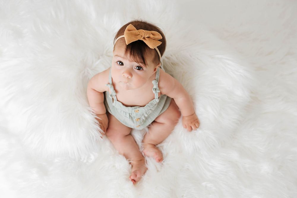 Adorable baby girl sitting on white fur rug wearing sage romper and gold bow headband.