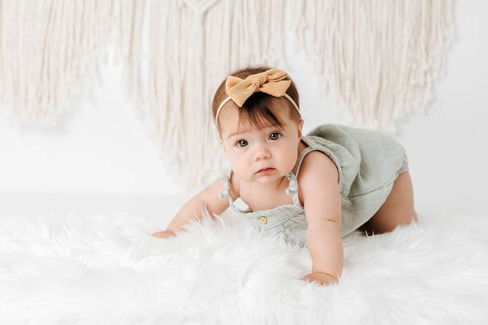 Cute baby girl in sage romper and tan bow headband crawling on white fluffy rug.
