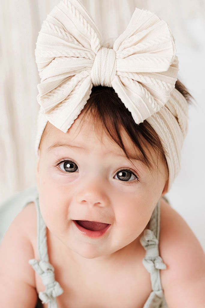 Smiling baby girl with big brown eyes wearing cream ribbed bow headband and sage romper.