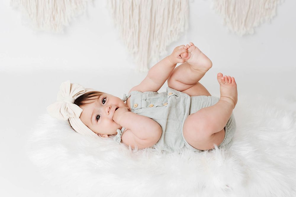 Adorable baby girl in sage romper lying on fluffy white rug, playfully holding her feet up in the air.