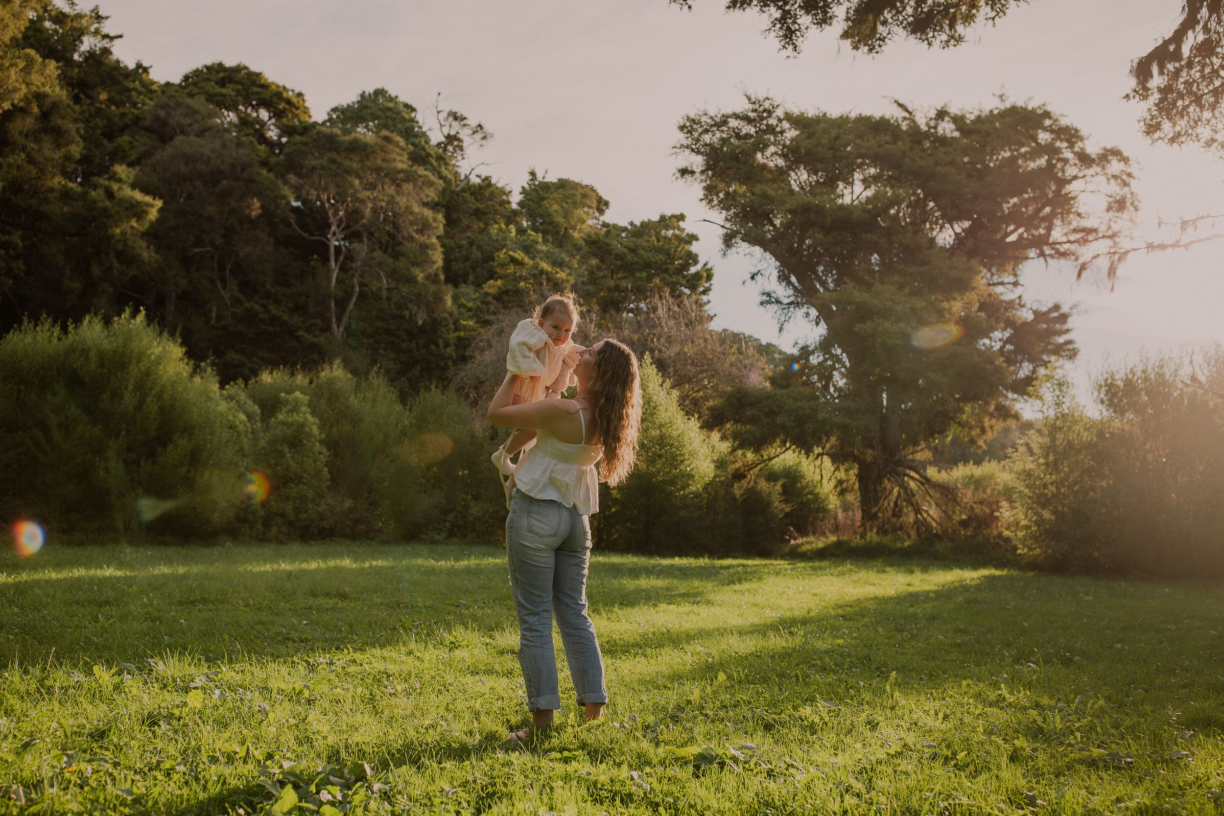 Mum and daughter, sunset in trees, Baigents reserve, motherhood photo session playing. Wide shot.