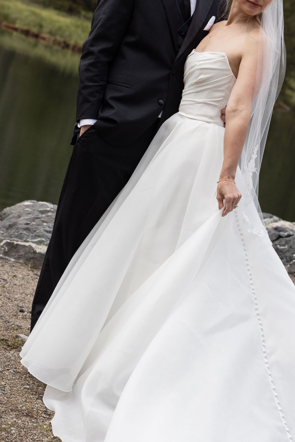 Bride in strapless white ball gown with veil stands beside groom in black tuxedo near a serene pond.