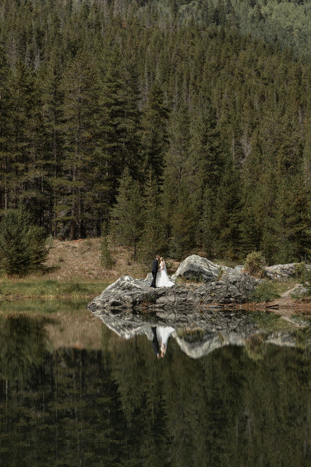 Bride and groom standing on rocks by a reflective mountain lake surrounded by evergreen forest.